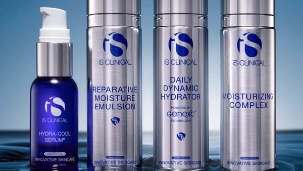 Four skincare bottles from iS Clinical arranged on a reflective dark surface against a blue gradient background. A dark blue pump bottle labeled 'Hydra-Cool Serum' stands on the left, while three tall silver bottles with blue labels read 'Reparative Moisture Emulsion,' 'Daily Dynamic Hydrator,' and 'Moisturizing Complex.