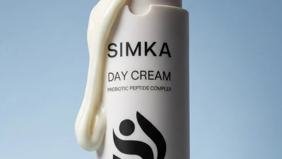 A 50ml bottle of Simka Day Cream SPF 15, featuring a white cylindrical container with black text and a black logo, with a thick swirl of off-white cream dripping from the cap against a soft blue gradient background.