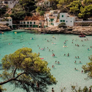 A scenic coastal cove with turquoise water filled with swimmers, white buildings, and green trees on a hillside under a pale sky.