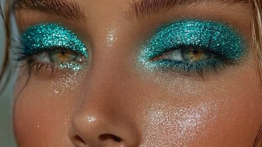 Close-up of a person with glittery teal eye makeup and pink lips.